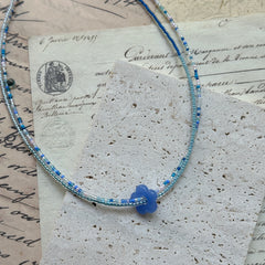 Azure Bloom Necklace close-up on rustic paper, featuring petite azure glass flower pendant with delicate strands of sky blue and pearl white beads