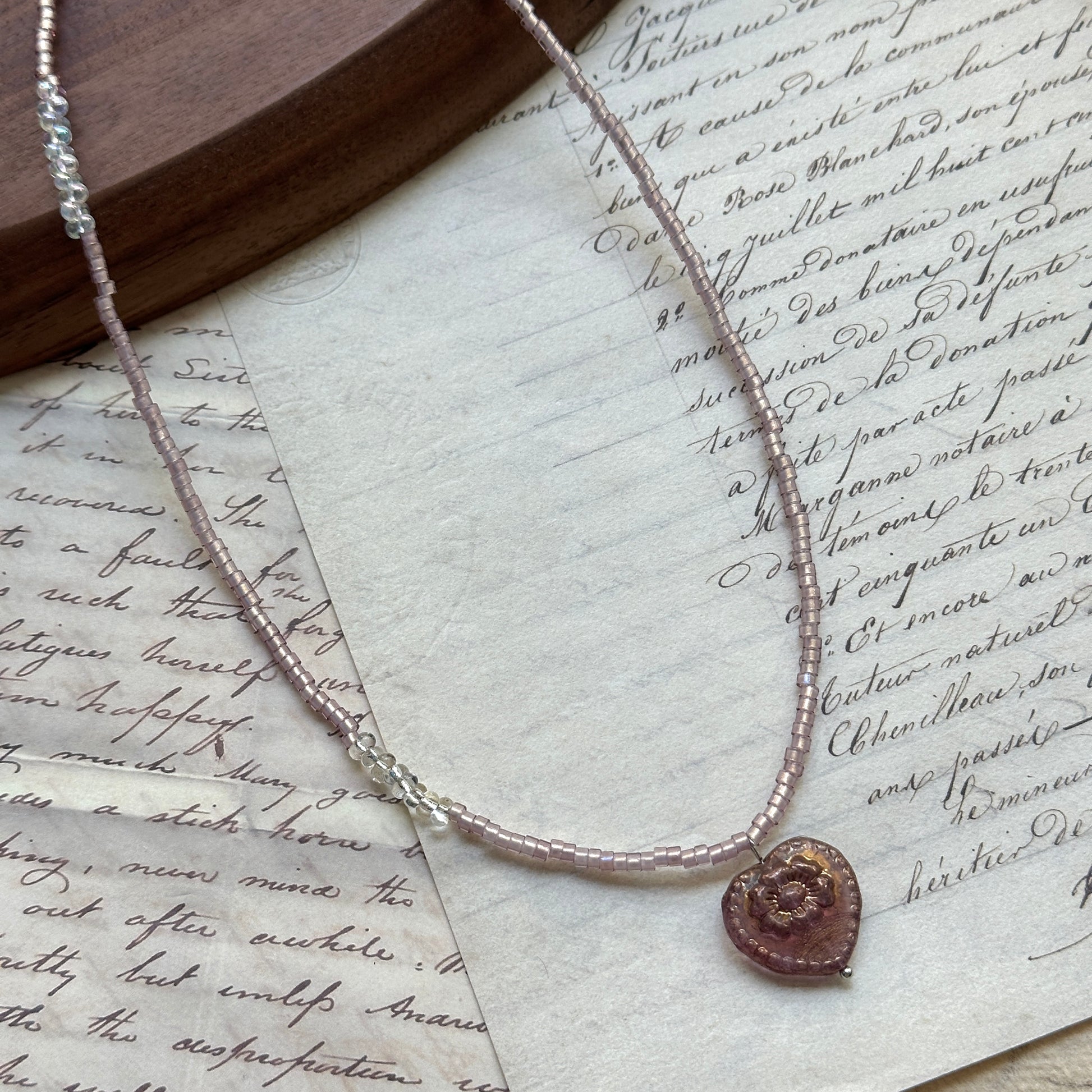 Pink Serenade Necklace close-up on rustic paper, featuring blush-pink seed beads with a luminous heart pendant and golden accents