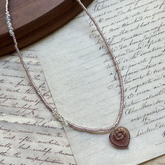 Pink Serenade Necklace close-up on rustic paper, featuring blush-pink seed beads with a luminous heart pendant and golden accents