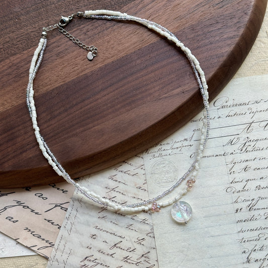 Opal Dream Necklace close-up on rustic paper, featuring double-layer white and blush seed beads with luminous opal pendant