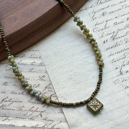 Gilded Bloom Necklace close-up on rustic paper, featuring golden and dark seed beads with engraved floral square charm