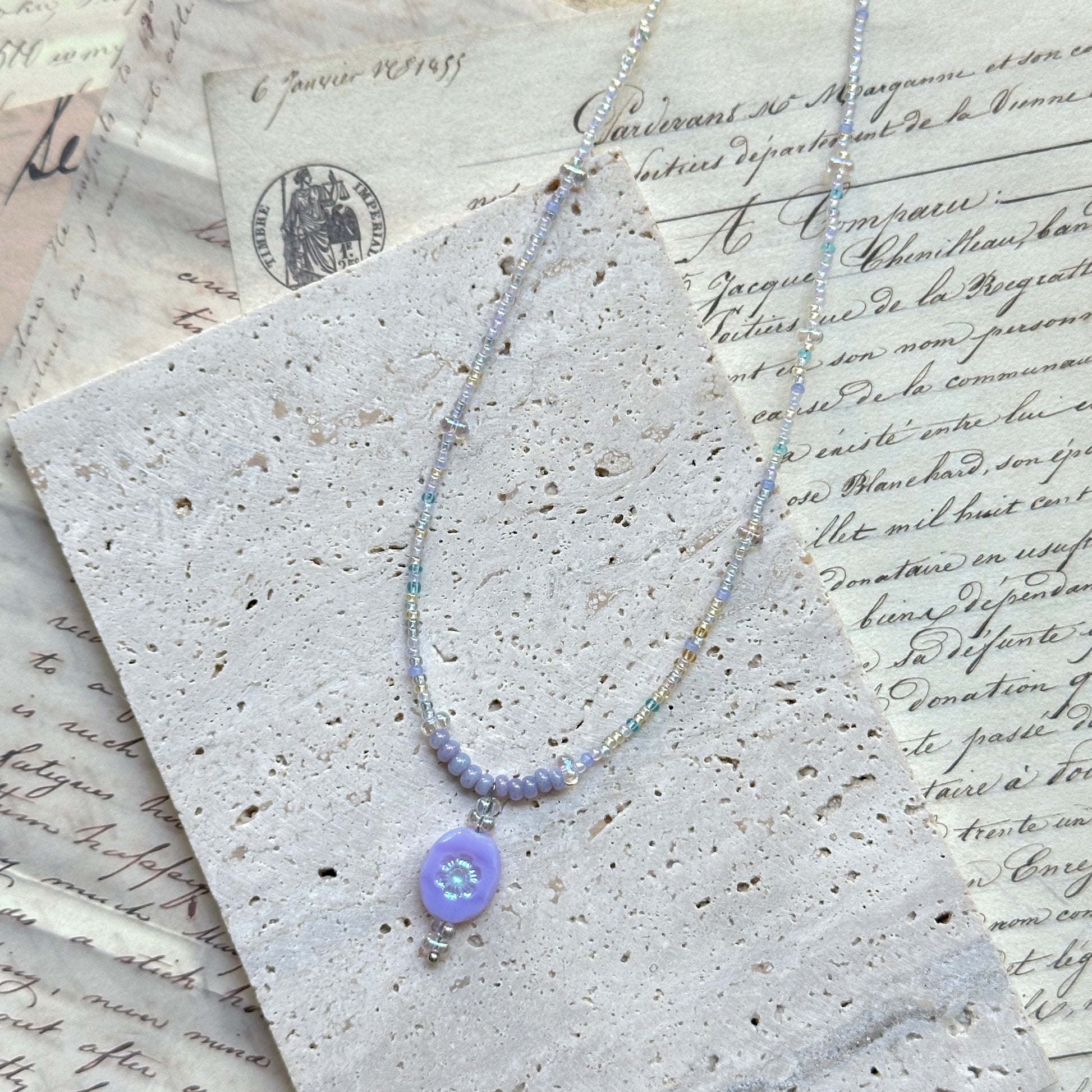 Aurora Dew Necklace flat lay on textured paper, featuring iridescent seed beads with lavender pendant and crystal drop