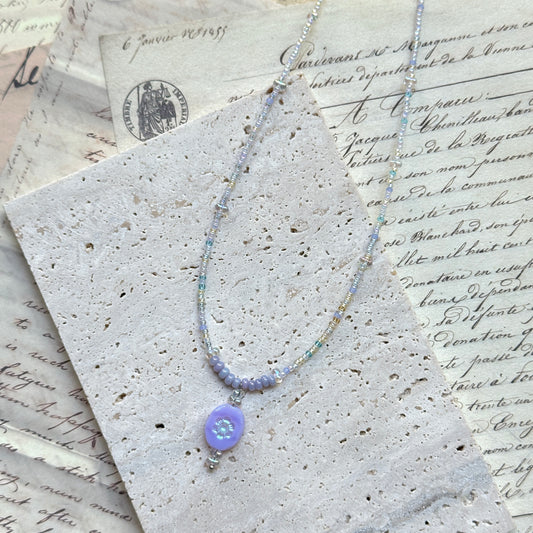 Aurora Dew Necklace flat lay on textured paper, featuring iridescent seed beads with lavender pendant and crystal drop