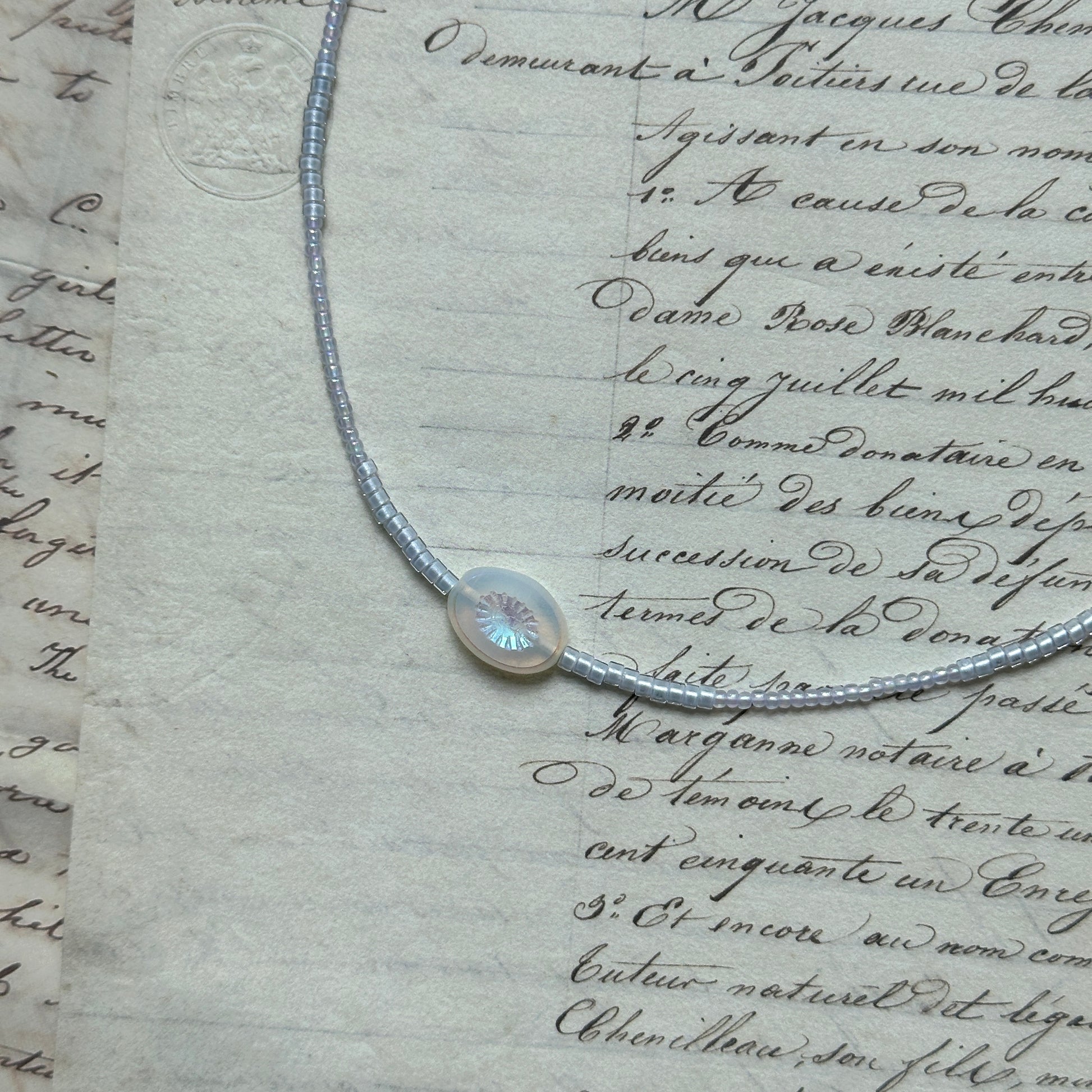 Dawn Halo Necklace flat lay on textured paper, featuring white seed beads with engraved round sun pendant