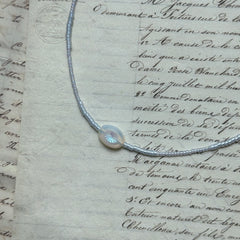 Dawn Halo Necklace flat lay on textured paper, featuring white seed beads with engraved round sun pendant