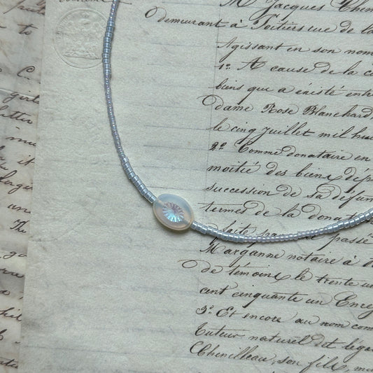 Dawn Halo Necklace flat lay on textured paper, featuring white seed beads with engraved round sun pendant