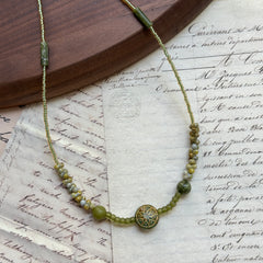 Verdant Bloom Necklace with gradient green and gold seed beads, floral medallion centerpiece, displayed on vintage paper background