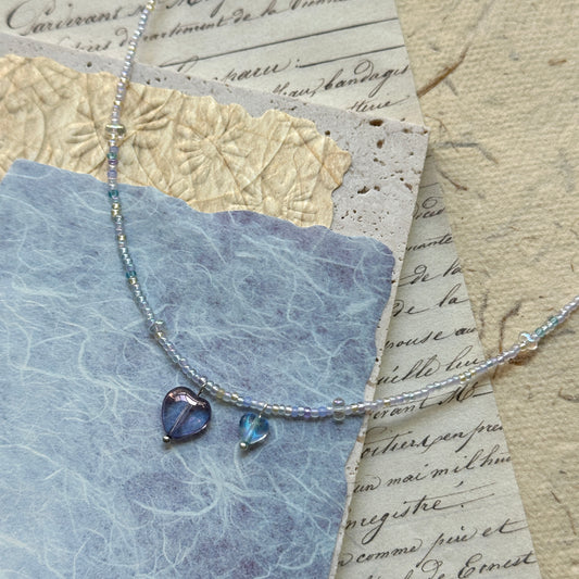 Celestial Love Necklace with violet glass heart charm and luminous blue bead on textured paper background