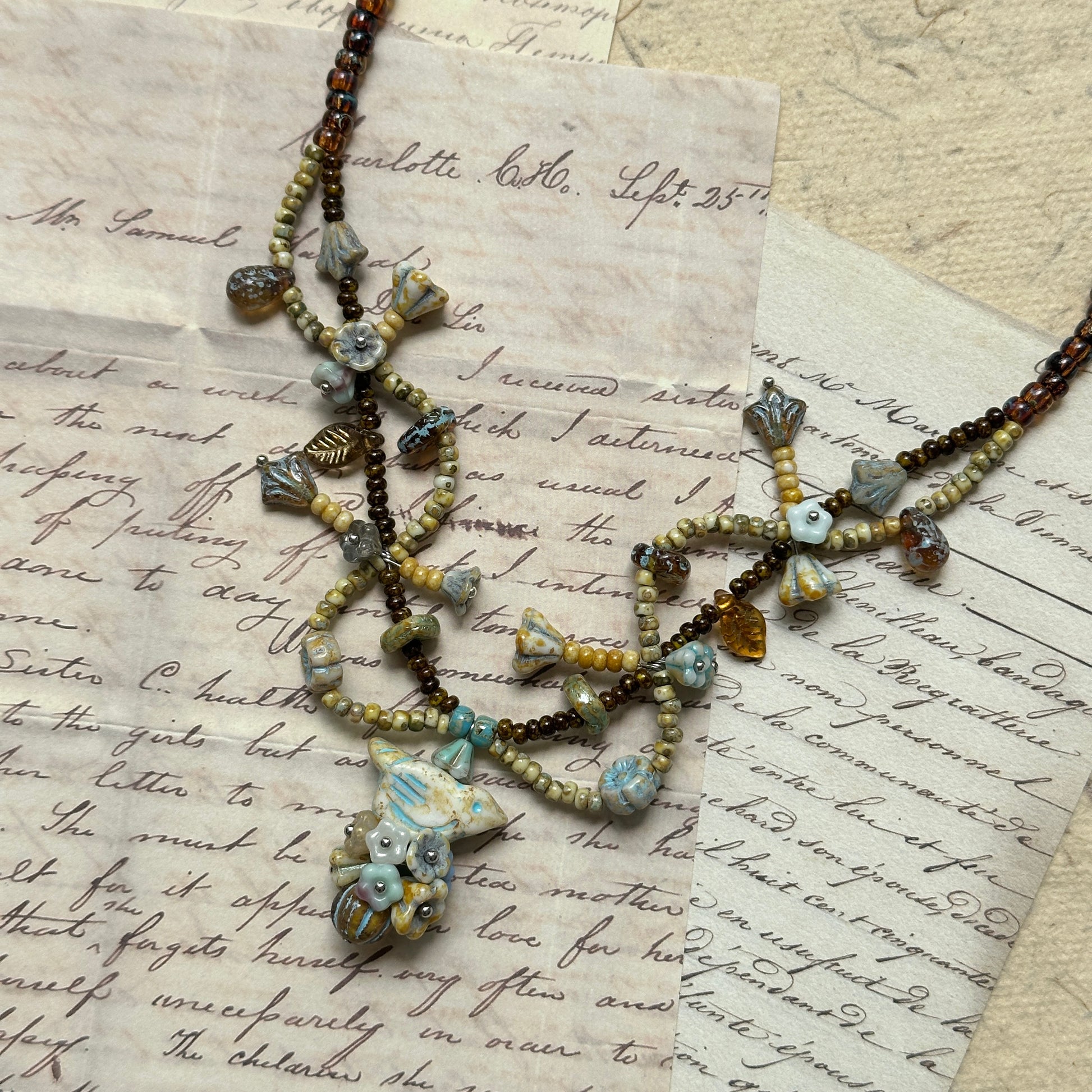 Autumn Whisper Necklace with golden-brown and soft blue tones, floral and leaf-shaped accents, shown on wooden necklace display stand.