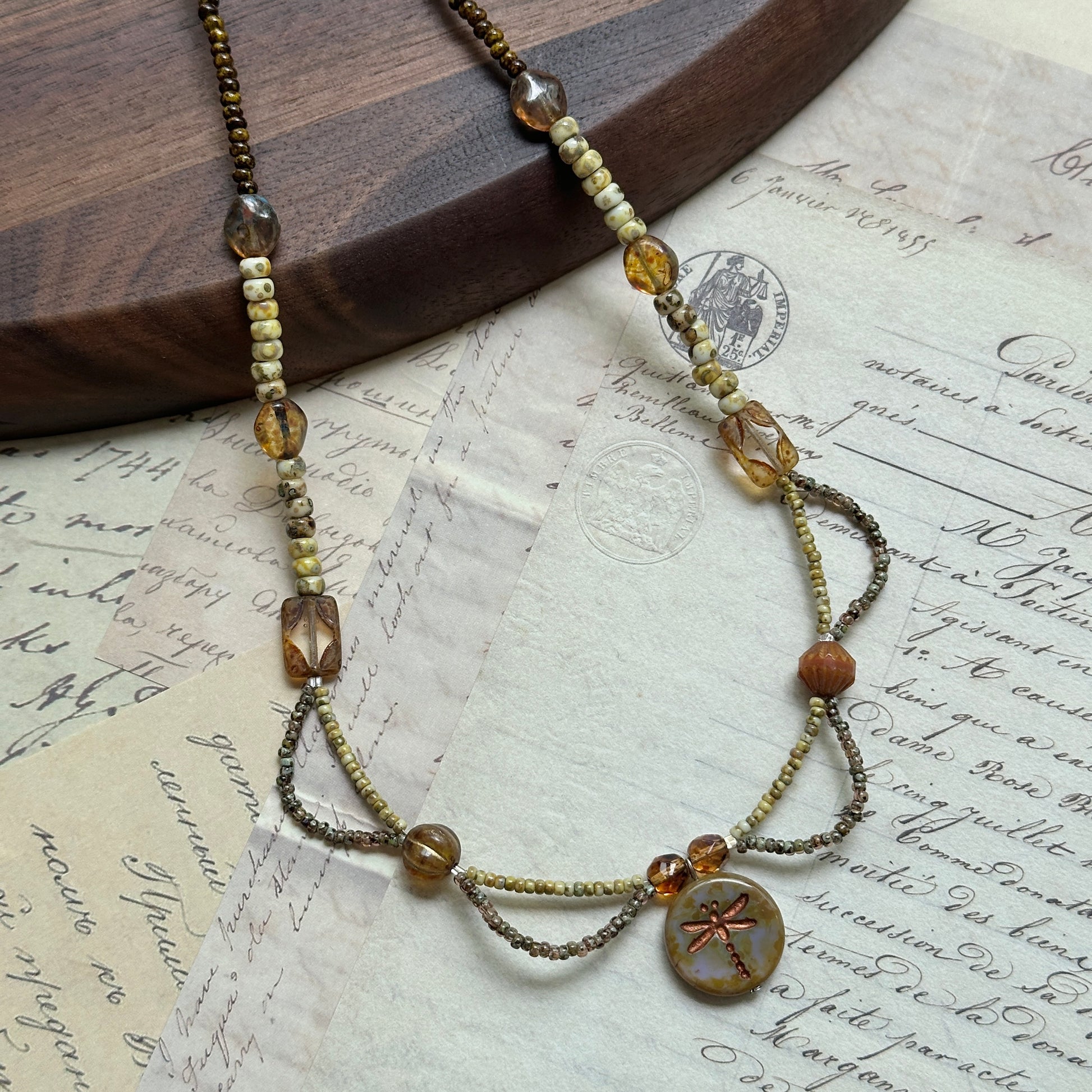Handcrafted Dragonfly Whisper Necklace with amber, earthy green, and warm brown beads, featuring a dragonfly medallion centerpiece, displayed on wooden background.