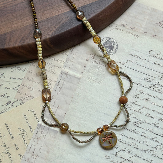 Handcrafted Dragonfly Whisper Necklace with amber, earthy green, and warm brown beads, featuring a dragonfly medallion centerpiece, displayed on wooden background.