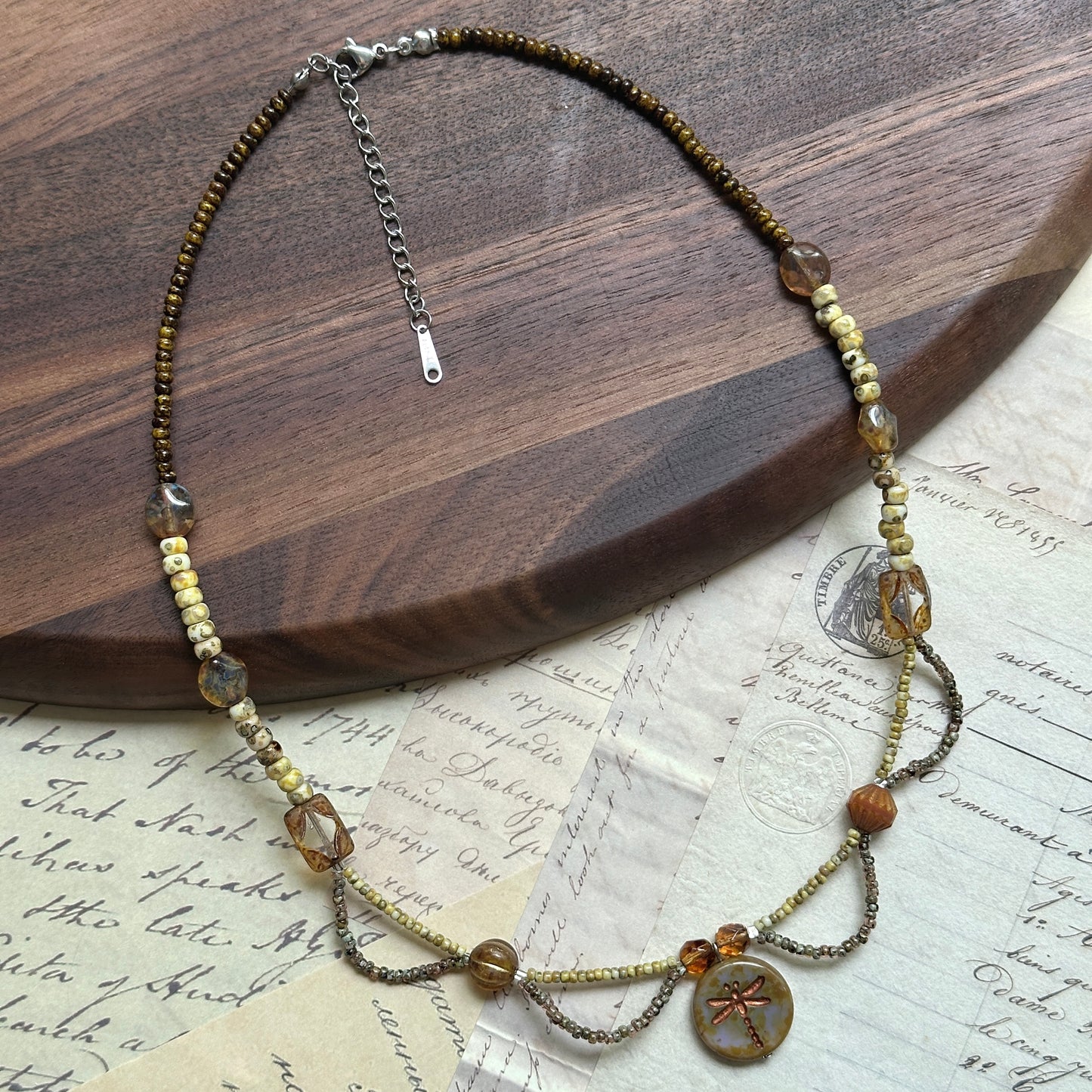 Artisan beaded necklace with dragonfly medallion, handcrafted using amber glass, Czech beads, and earthy green tones, arranged in circular display on white surface.
