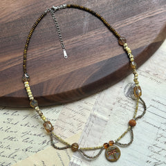 Artisan beaded necklace with dragonfly medallion, handcrafted using amber glass, Czech beads, and earthy green tones, arranged in circular display on white surface.
