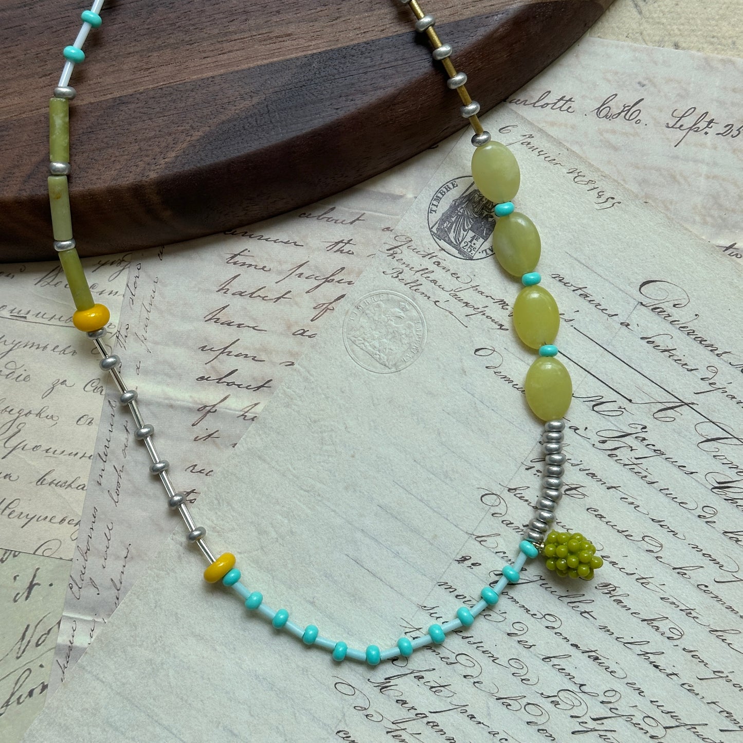 Asymmetrical artisan necklace combining turquoise, green, and yellow beads with a grape-cluster charm, arranged in circular display on white surface.