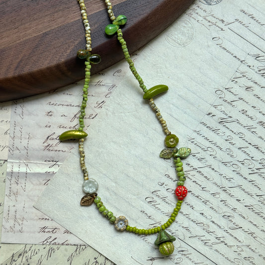 Handcrafted Toadstool Charm Necklace with green seed beads, woodland mushroom, leaf, flower, and red berry accents, displayed on wooden background.