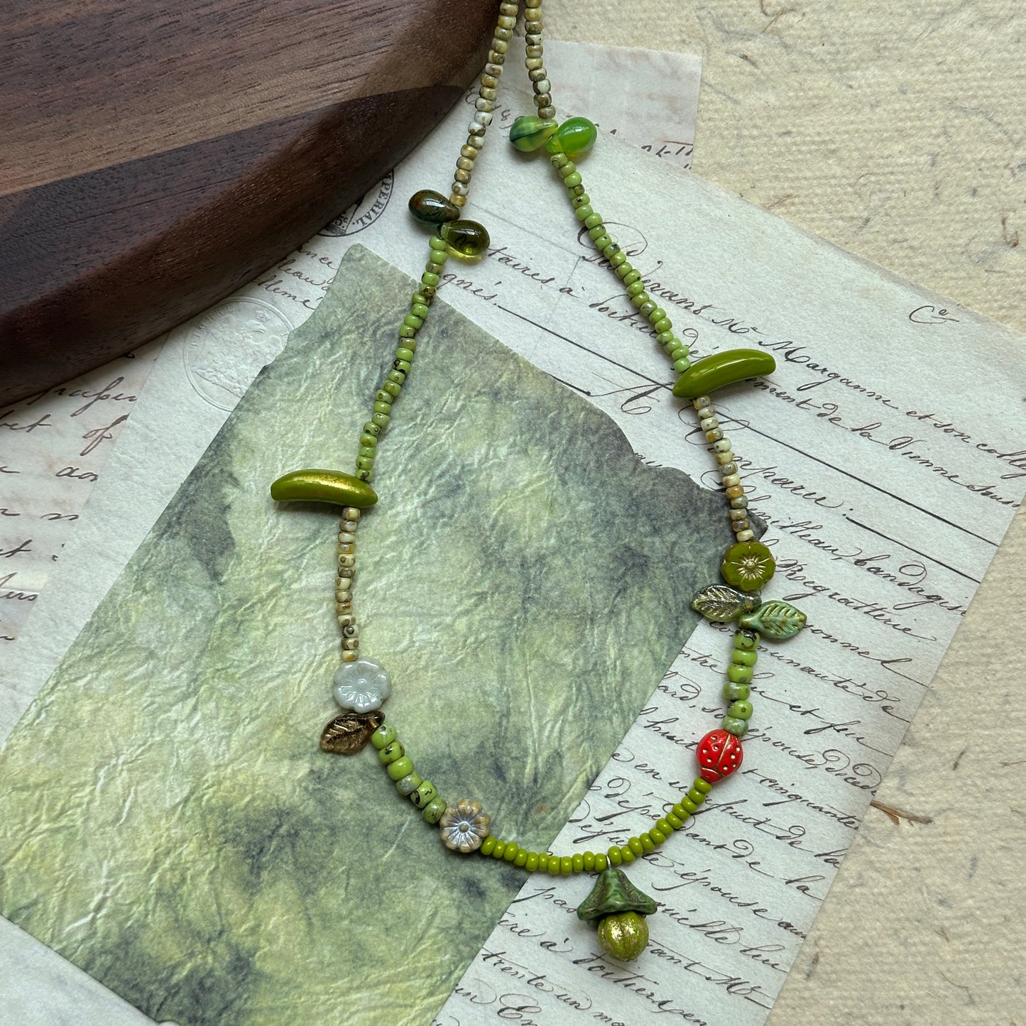 Whimsical artisan necklace featuring green seed beads with woodland-inspired charms—mushroom, leaves, flowers, and tiny red berry—arranged in circular display.