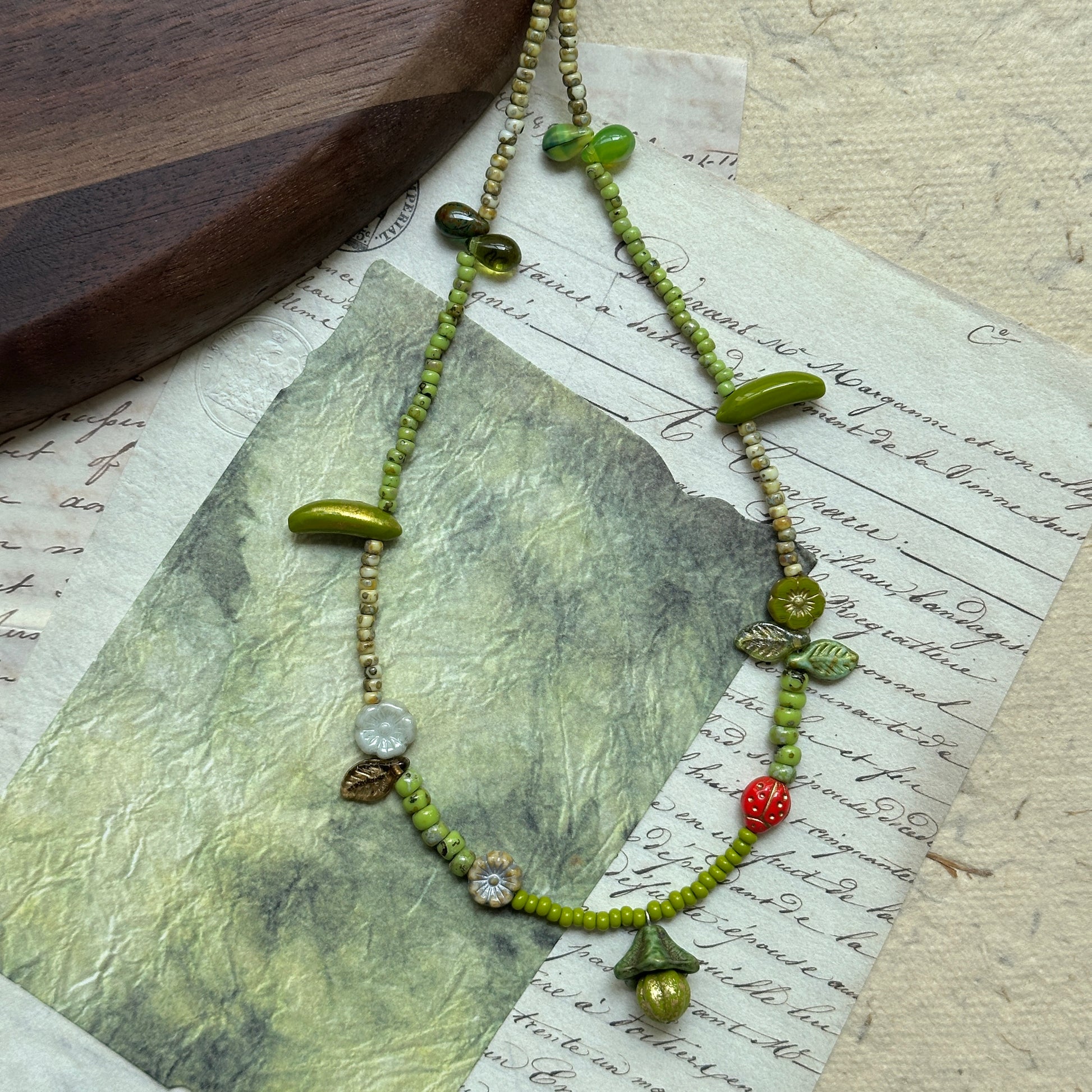 Whimsical artisan necklace featuring green seed beads with woodland-inspired charms—mushroom, leaves, flowers, and tiny red berry—arranged in circular display.