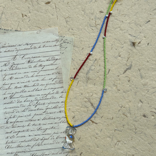 Handcrafted Joyful Hour Necklace with rainbow-colored seed beads and a crystal-like hourglass pendant, displayed flat on vintage paper background.