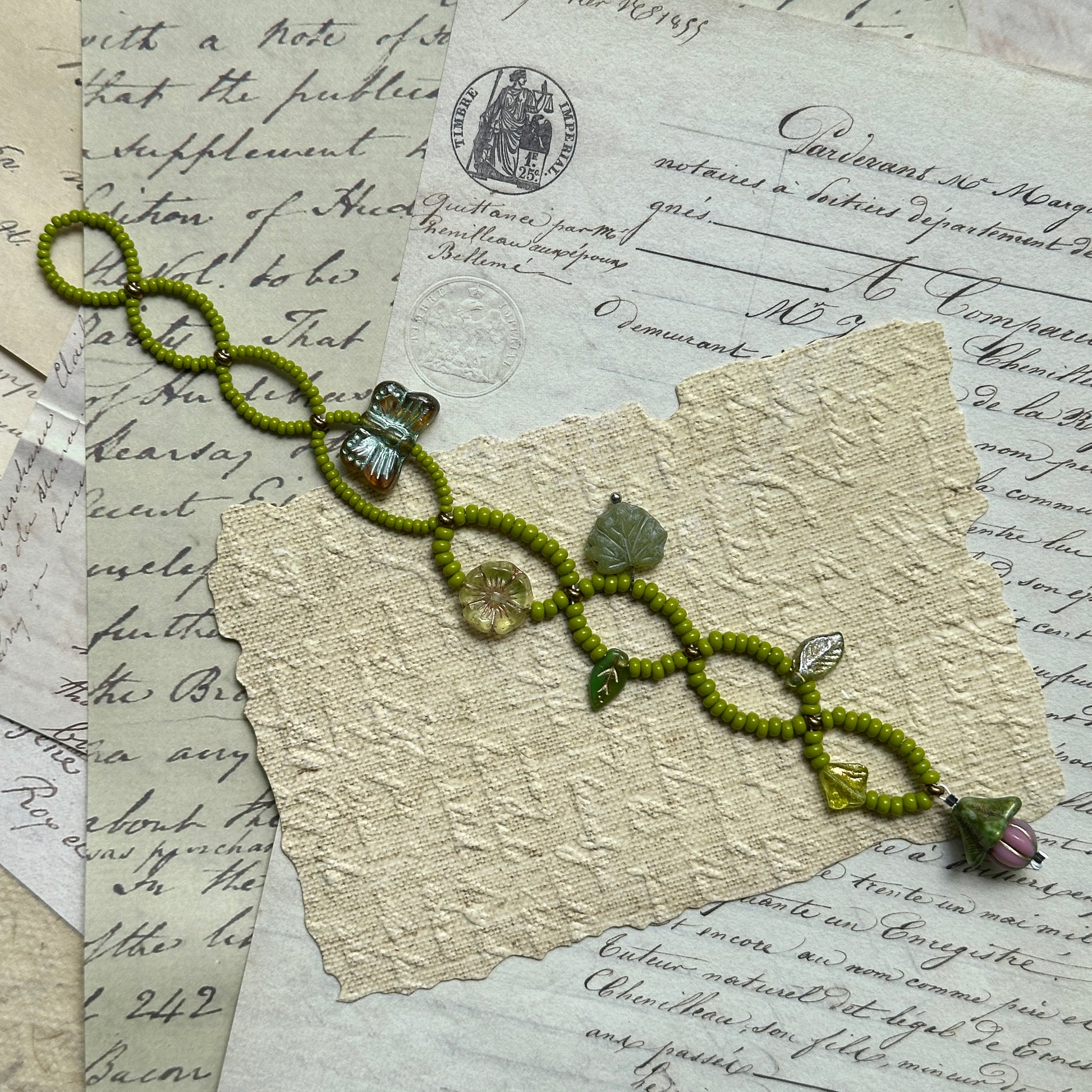 Handcrafted Meadow Charm Bracelet with fresh green beads, leaf accents, butterfly and flower motifs, displayed flat on vintage paper background.