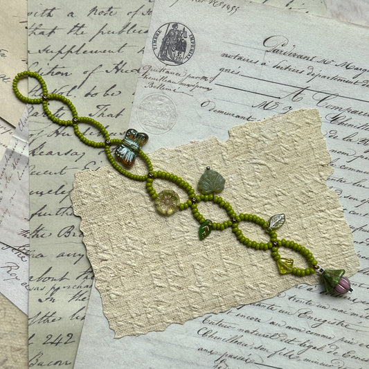 Handcrafted Meadow Charm Bracelet with fresh green beads, leaf accents, butterfly and flower motifs, displayed flat on vintage paper background.