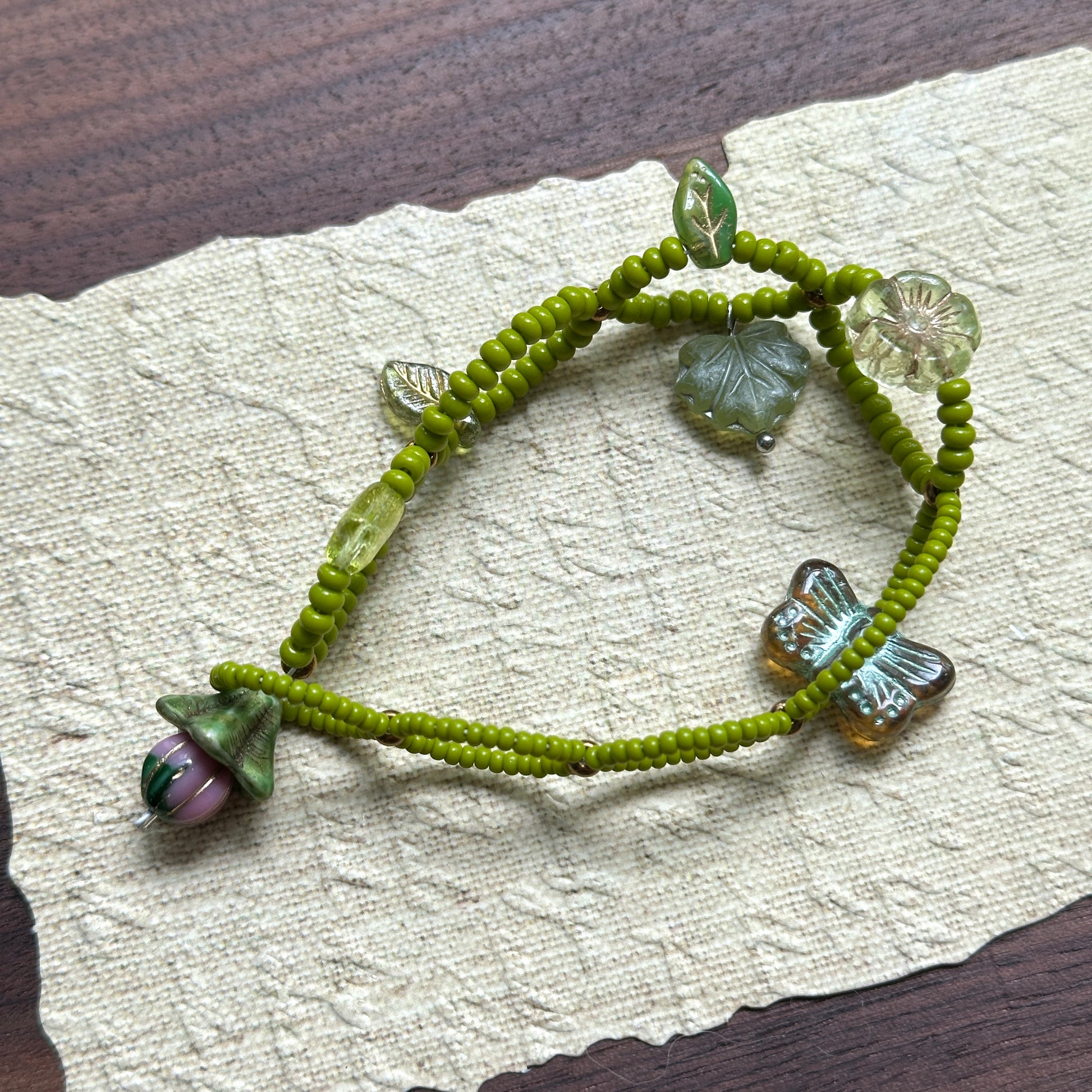 Artisan bracelet with whimsical woodland theme, featuring green beads, delicate leaf details, and a butterfly charm, arranged in circular display.