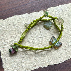 Artisan bracelet with whimsical woodland theme, featuring green beads, delicate leaf details, and a butterfly charm, arranged in circular display.