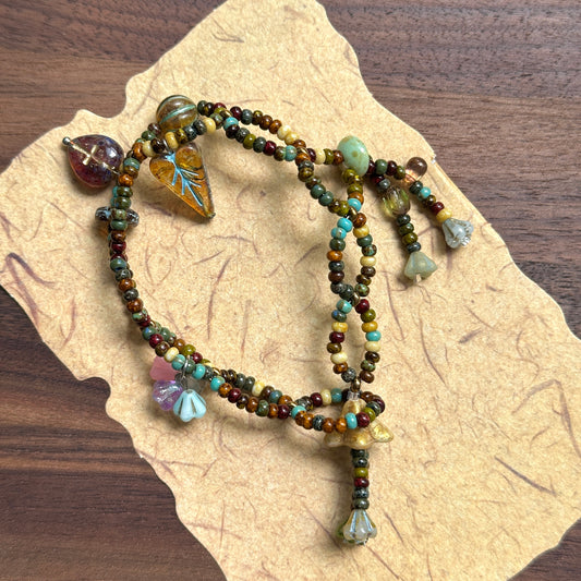 Handcrafted Harvest Charm Bracelet with amber, brown, and green beads, featuring leaf, flower, and bell-shaped charms, displayed flat on parchment background.