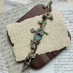 Handcrafted Blue Garden Bracelet with soft blue, beige, and golden-brown beads, featuring a central blue floral medallion and leaf accents, displayed flat on wooden surface.