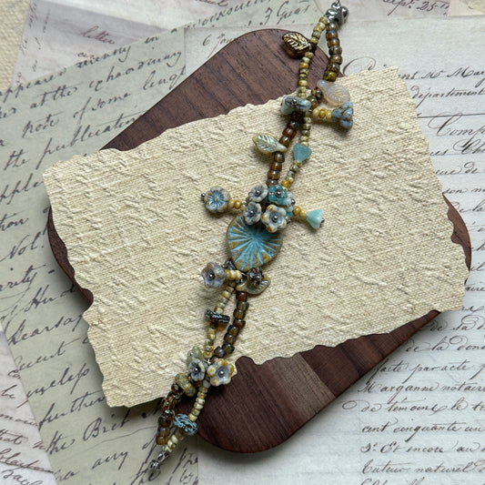 Handcrafted Blue Garden Bracelet with soft blue, beige, and golden-brown beads, featuring a central blue floral medallion and leaf accents, displayed flat on wooden surface.