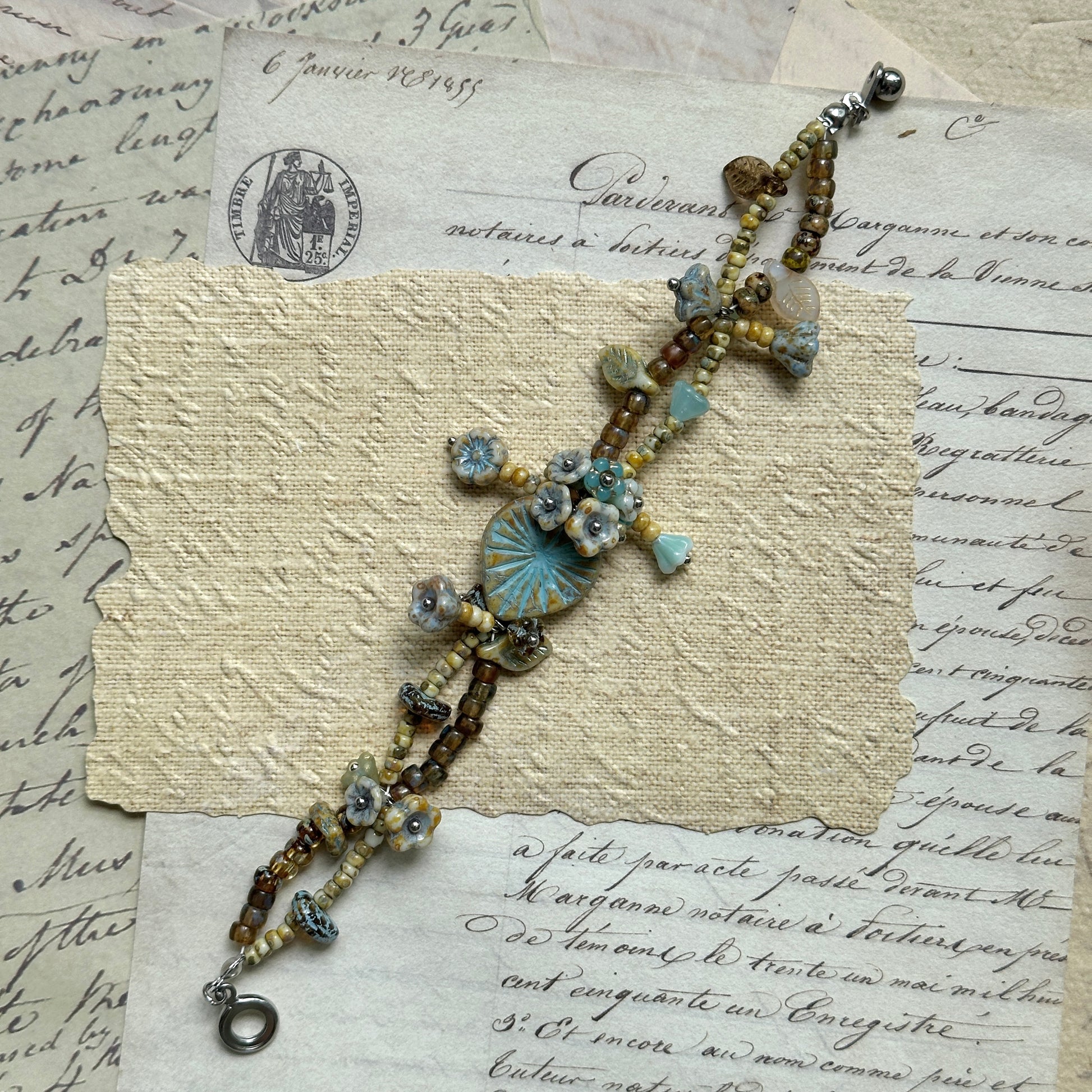 Artisan bracelet in sky blue, beige, and warm brown tones, decorated with floral medallion, tiny flowers, and leaf charms, arranged on vintage paper background.