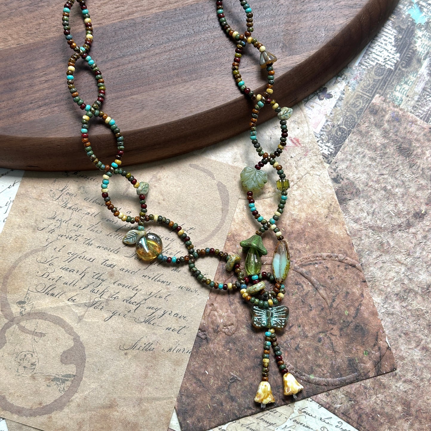 Olive Grove Lariat Necklace with Czech-glass leaves, hearts and a butterfly tassel in olive/moss/amber, flat lay on wood.