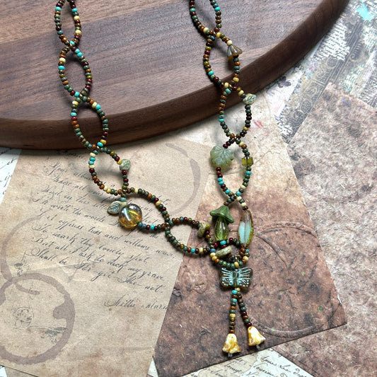 Olive Grove Lariat Necklace with Czech-glass leaves, hearts and a butterfly tassel in olive/moss/amber, flat lay on wood.