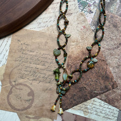 Angled flat lay of the Olive Grove Lariat Necklace showing full length, tassel accents and the woodland olive-amber palette.