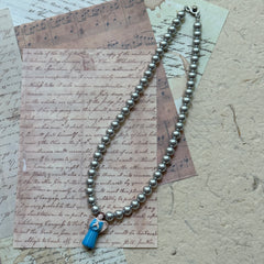 Angled flat lay of the Angel’s Grace Necklace—timeless faux pearls with a delicate blue angel charm.