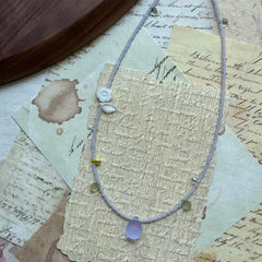 Lilac Dew Necklace coiled flat lay showing full strand with scattered pale-green and soft-yellow accents and a lilac teardrop focal.