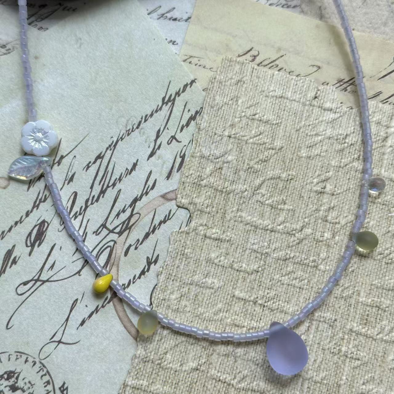 Angled flat lay on parchment background—minimal lavender bead strand with frosted lilac teardrop, serene spring look.