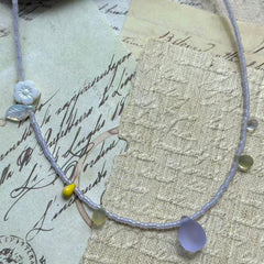 Angled flat lay on parchment background—minimal lavender bead strand with frosted lilac teardrop, serene spring look.