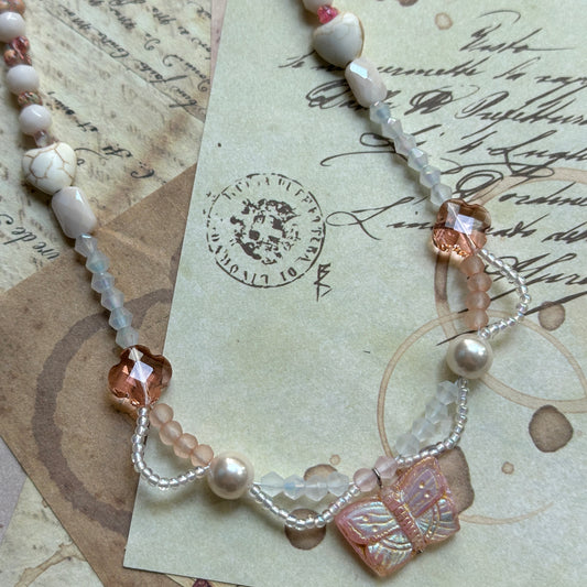 Close detail of Blush Wing Necklace—blush Czech-glass butterfly, crystal flower charms, seed beads and faux-pearl accents; flat lay.