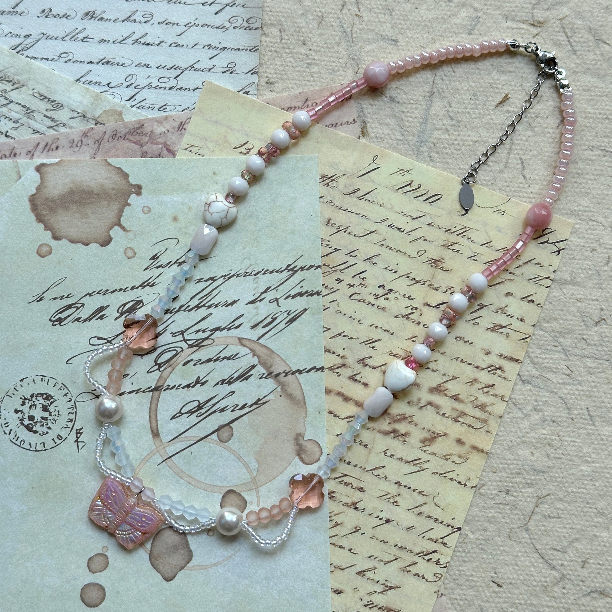 Pendant close-up on vintage paper—rose-pink Czech-glass butterfly with ivory seed-bead swags, faceted crystals and pearl accents.
