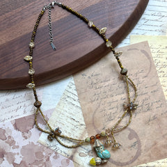 Angled flat lay on parchment—botanical necklace with Czech-glass flower charms and amber beads surrounding a turquoise leaf focal.