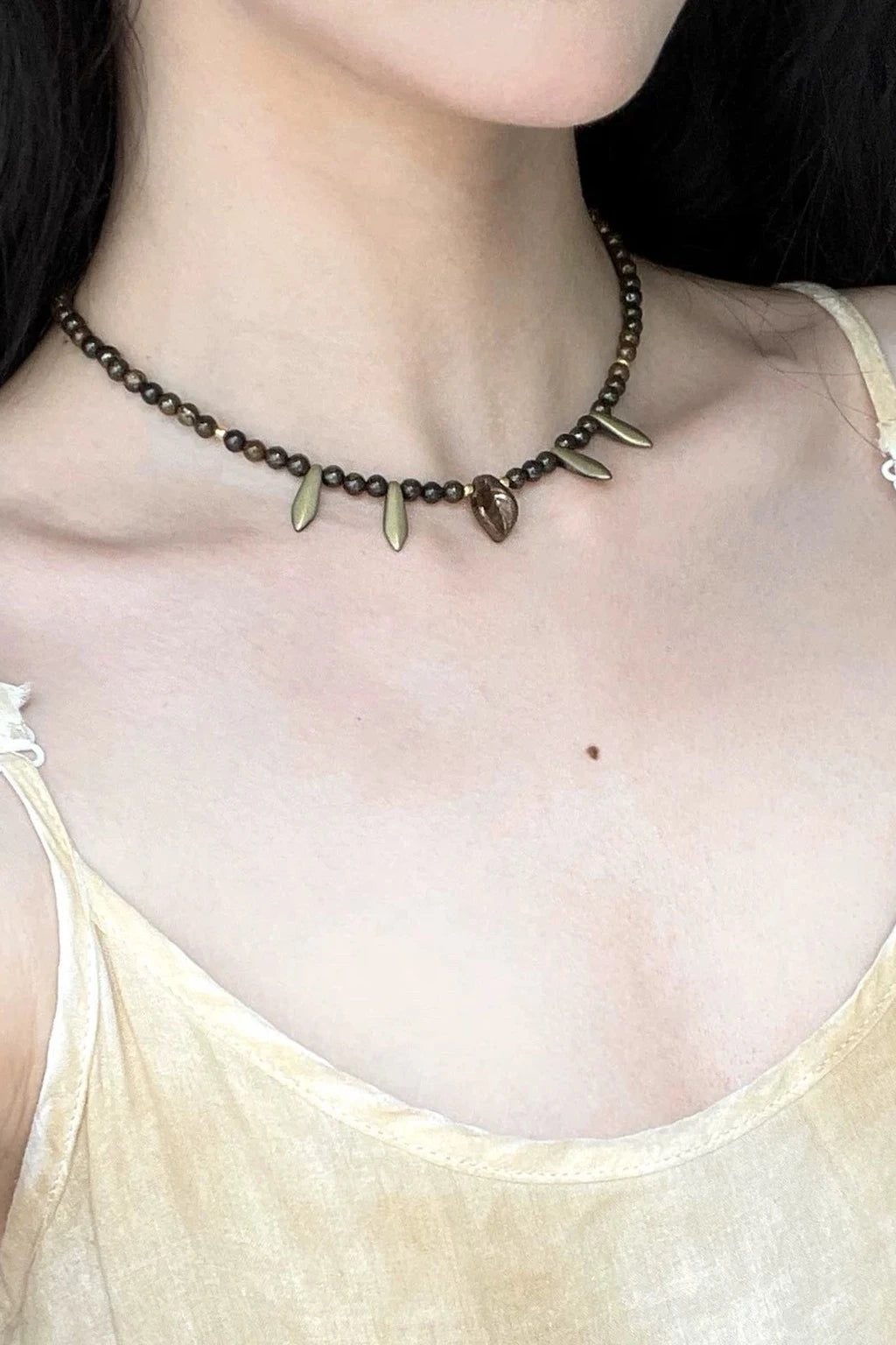 wearing a dark beaded choker necklace with small gold-toned charms, including leaf and fang-shaped pendants, styled with a light beige camisole in natural light.