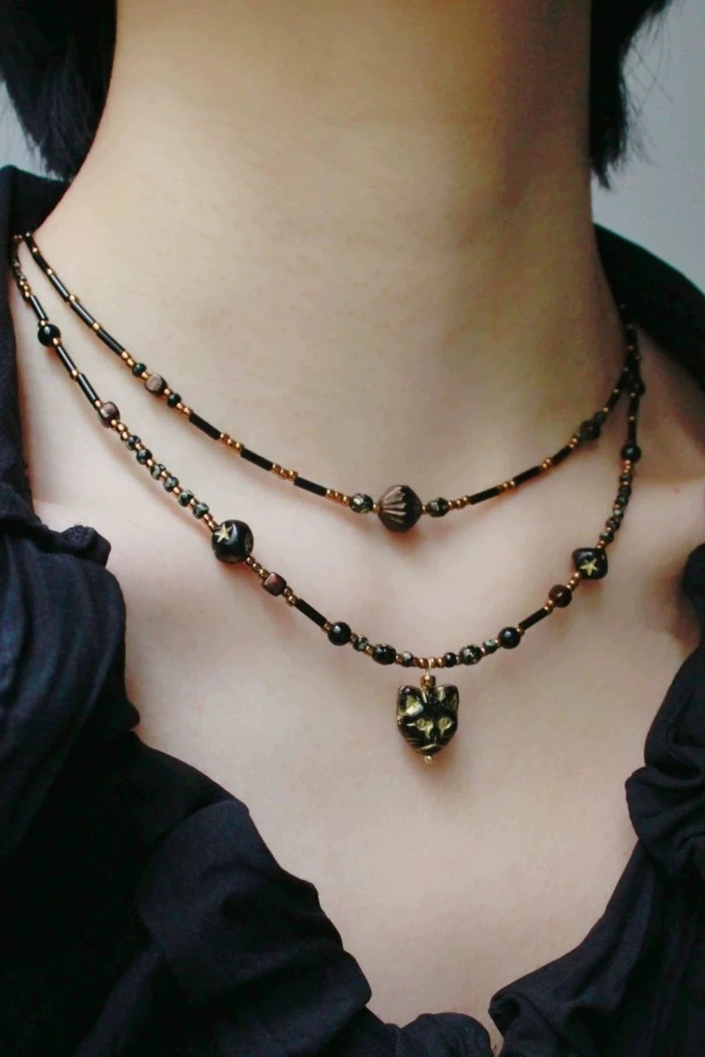 wearing layered dark beaded chokers in black and amber tones with gold accents, featuring a small cat-face heart pendant, styled with a black ruffled top.