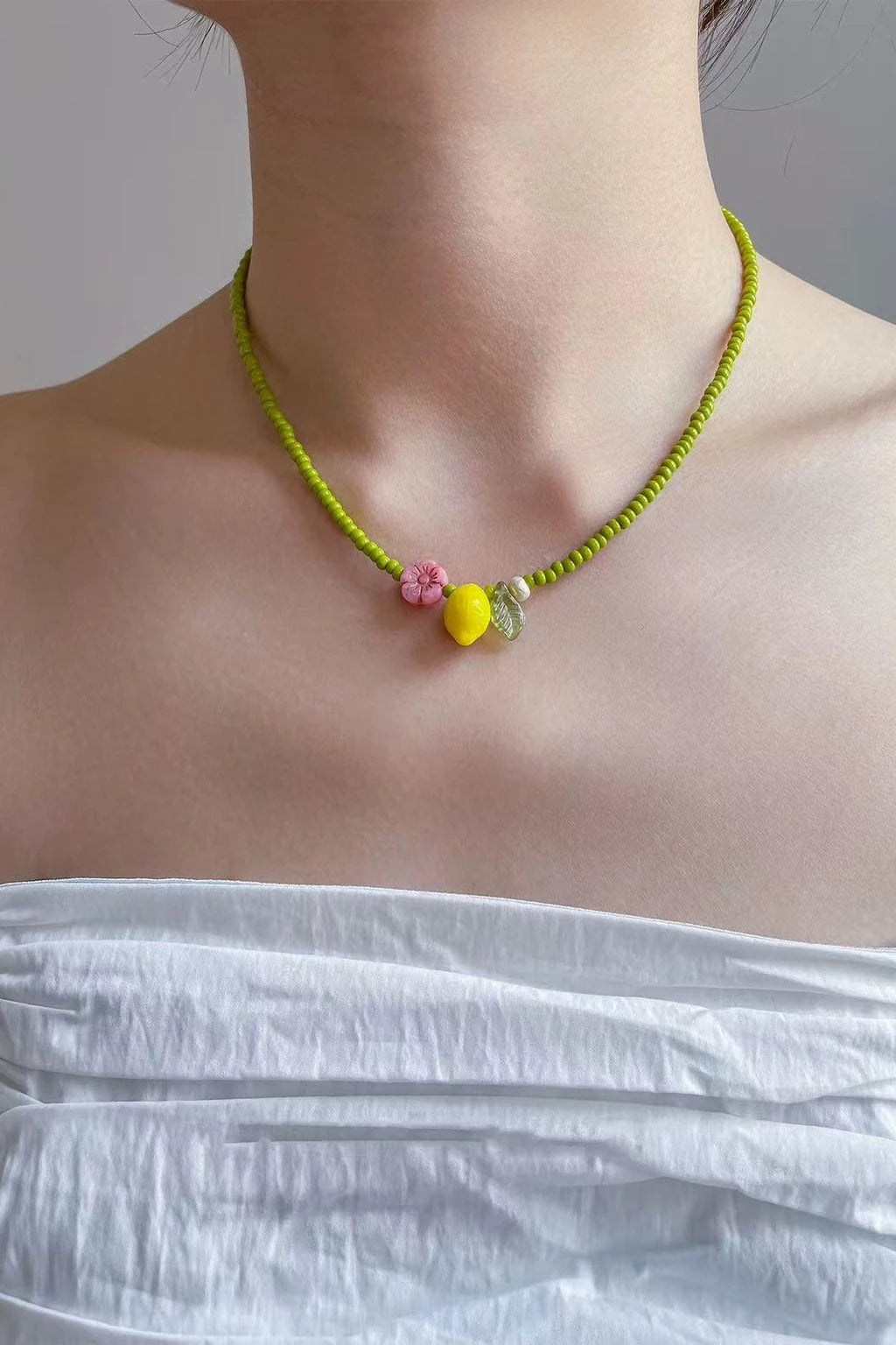 wearing a lime-green seed-bead necklace with a pink flower charm, yellow drop bead, and clear nugget, shown against a neutral background with a white off-shoulder top.