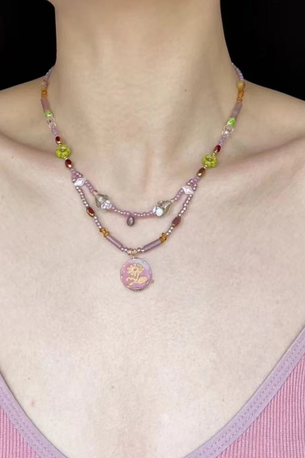 wearing layered pastel beaded necklaces in pink and lilac tones with mixed glass/Czech beads and a round floral pendant, shown against a black background and pink V-neck top.