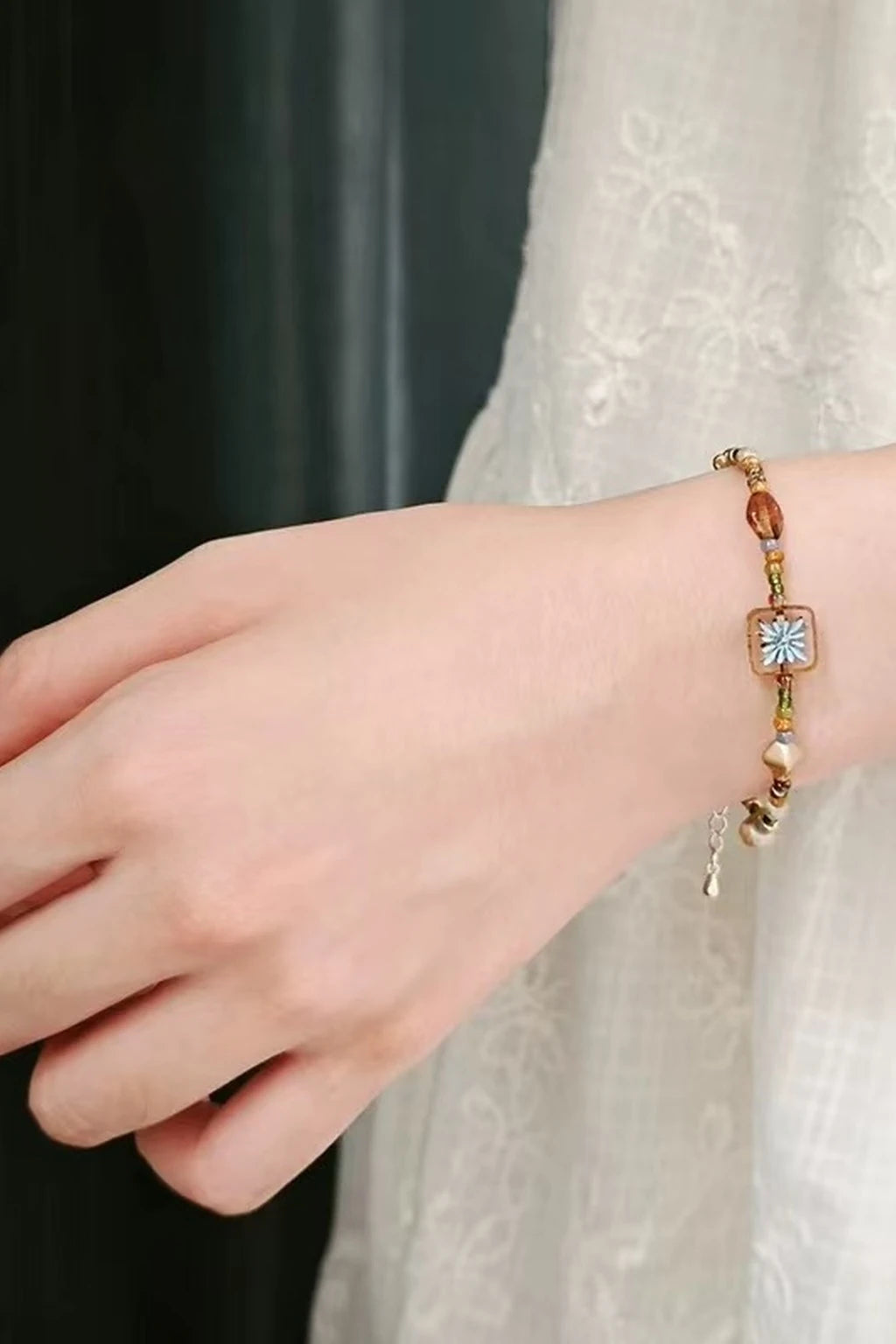 wrist wearing a vintage-style beaded bracelet with a small square floral tile bead (blue center), amber/gold beads, and pearl accents, photographed beside a white lace sleeve.
