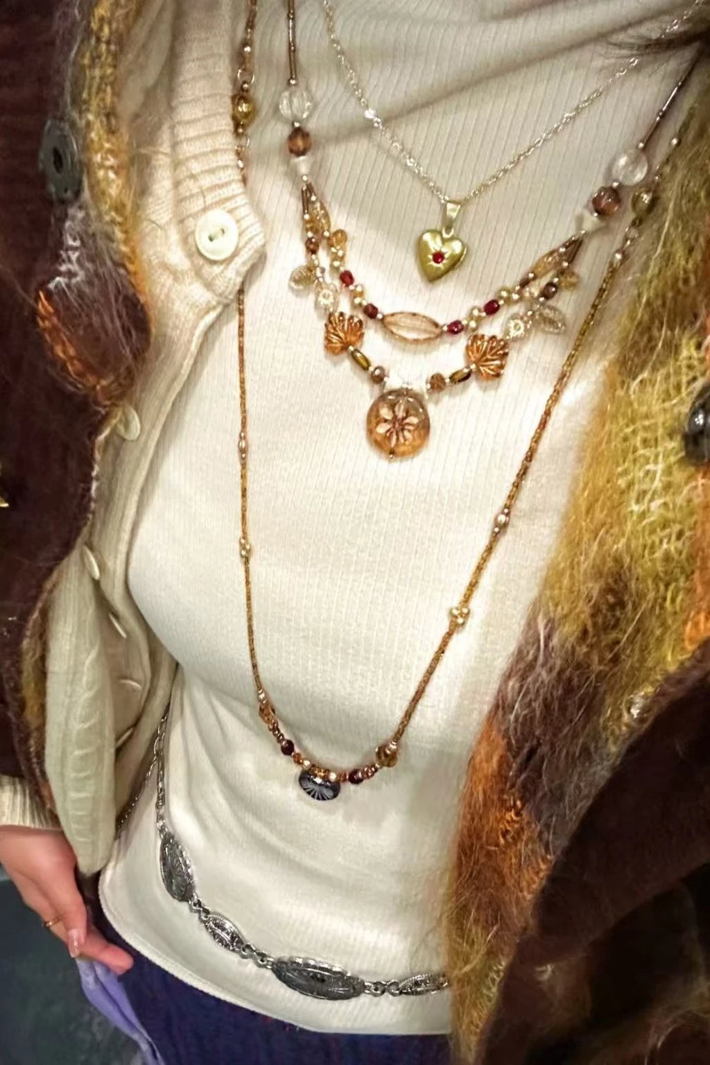 wearing layered necklaces: gold heart pendant, handcrafted beaded designs with amber and crystal tones, and a long chain with accents, styled over a cream turtleneck and cardigan.