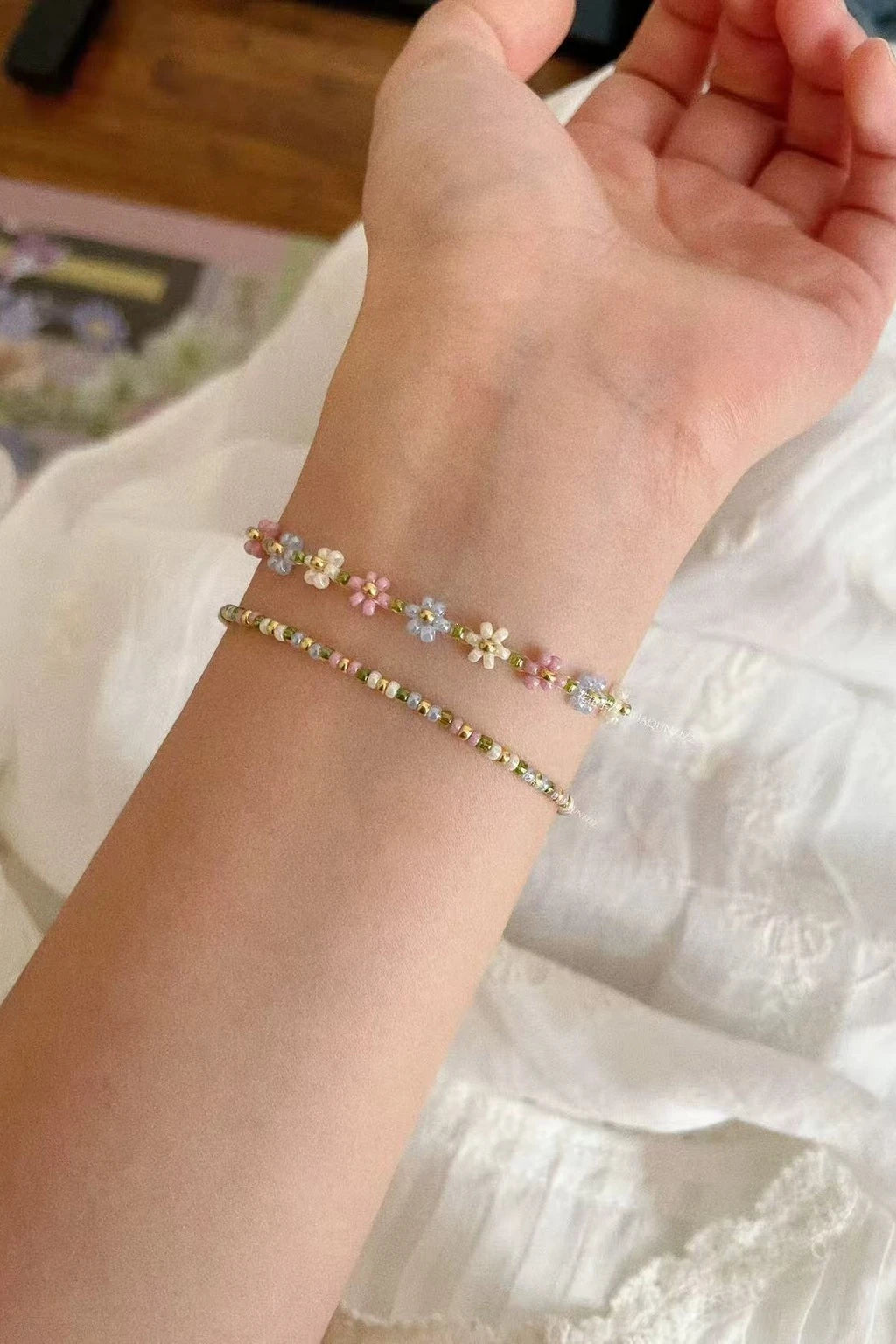 wrist wearing two handcrafted bracelets—a pastel daisy-chain and a slim seed-bead strand—photographed on white fabric in soft light.