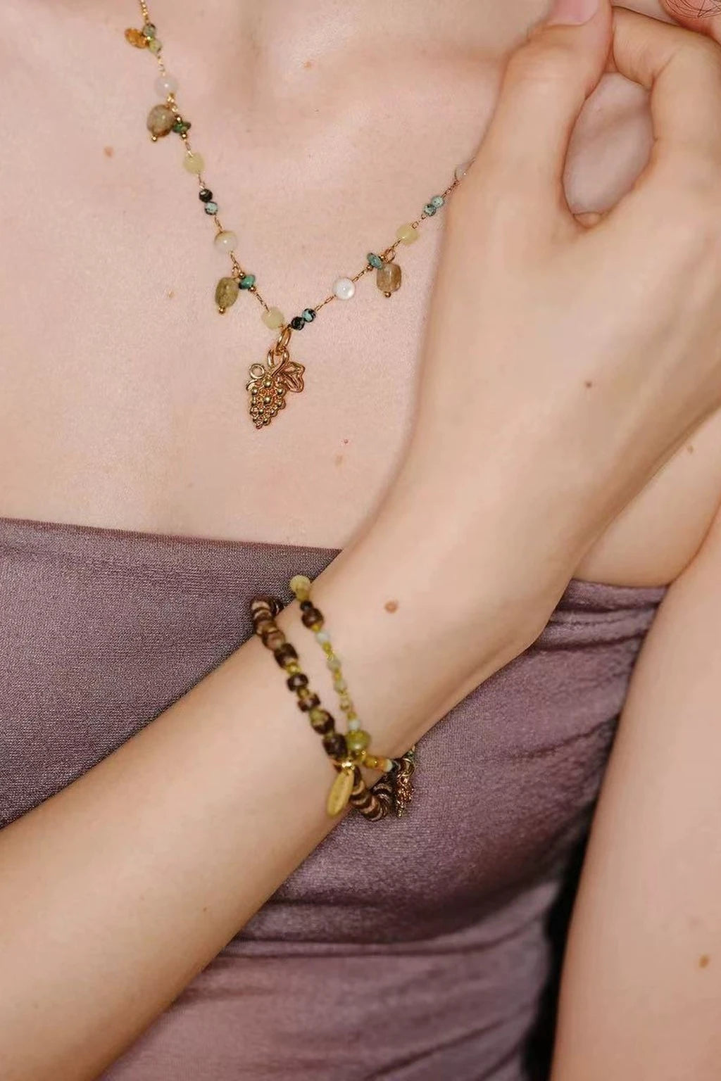 wearing an olive-amber beaded necklace with a small grape pendant and matching layered bracelets, photographed against a mauve strapless top with hand near the collarbone.
