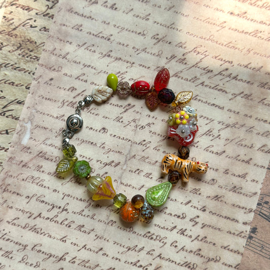 Tiger Orchard Bracelet handcrafted with a playful tiger bead, fruits, leaves, and amber-toned glass charms, displayed on vintage script paper background