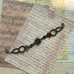 Antique Flower Window Bracelet with central floral medallion, amber and garnet Czech glass beads, bronze accents, flat-lay on vintage script paper